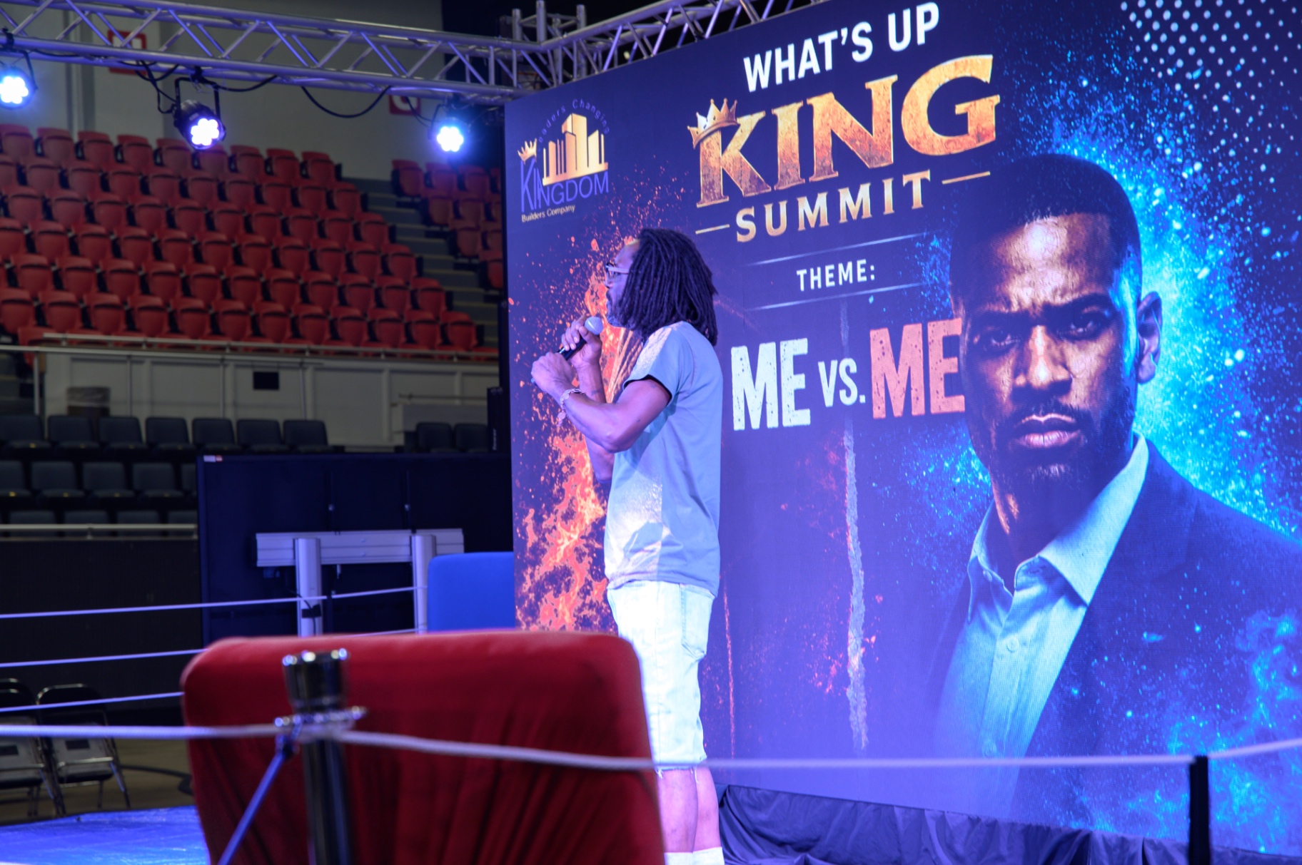 A summit speaker addressing the audience beside the illuminated Me vs. Me display